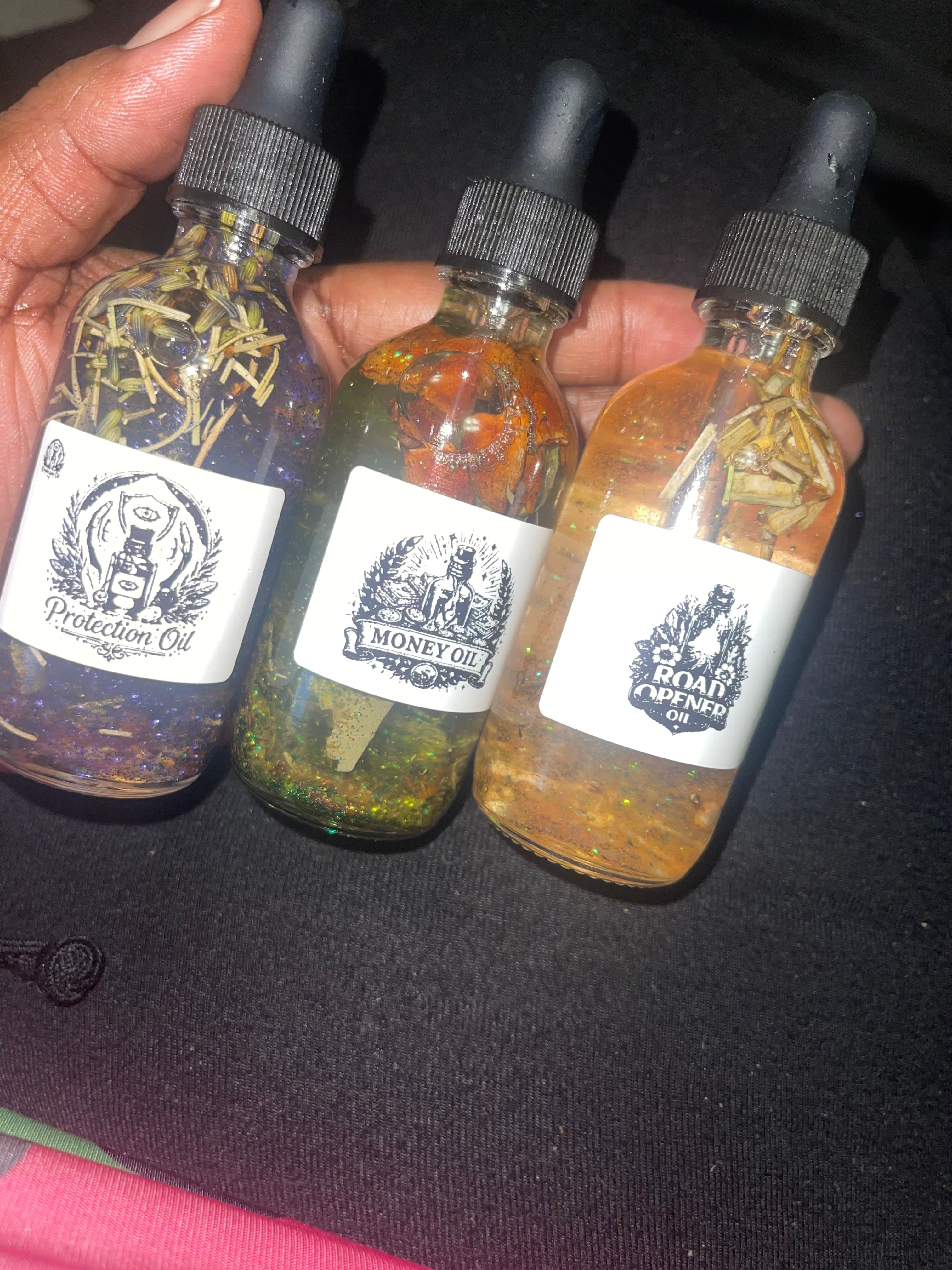 Hand holding three glass dropper bottles labeled Protection Oil, Money Oil, and Road Opener Oil.